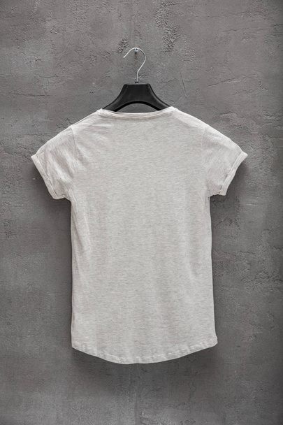 Back side of female grey melange cotton t-shirt on a hanger and a concrete wall in the background. T-shirt without print - Foto, afbeelding