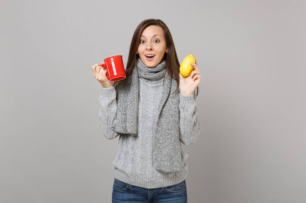 グレーのセーターに興奮した若い女性は、灰色の背景に分離された紅茶のレモンと赤のカップを保持しているマフラーします。健全なライフ スタイル、人々 の心から感情、寒い季節の概念。コピー スペースをモックアップします。 - 写真・画像