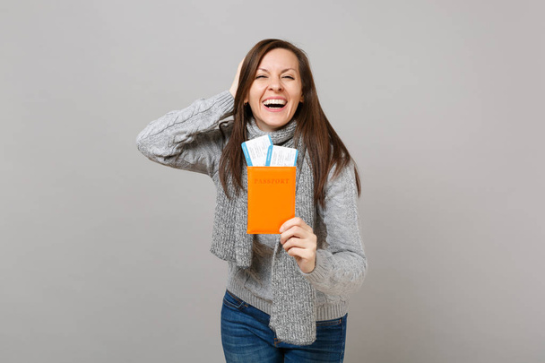 Śmiejąc się młoda kobieta w szary sweter, szalik kładą rękę na głowie, posiadający paszport boarding pass bilet na białym tle na szarym tle. Moda na zdrowy styl życia osób szczere emocje, koncepcja zimnej pory roku - Zdjęcie, obraz