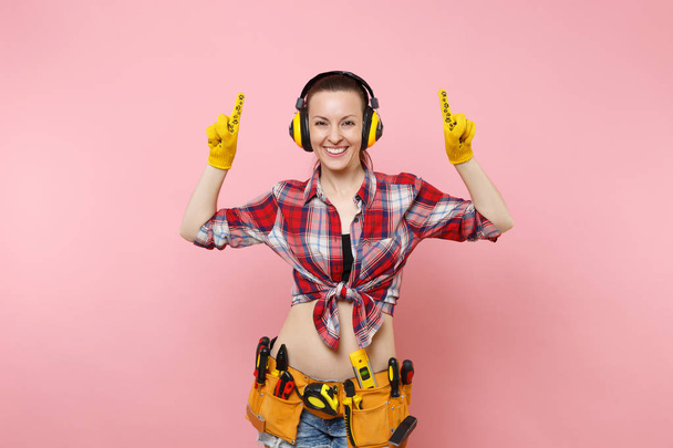 Handyman woman in plaid shirt, denim shorts, yellow gloves, noise insulated headphones, kit tools belt full of variety instruments isolated on pink background. Female in male work. Renovation concept - Photo, Image