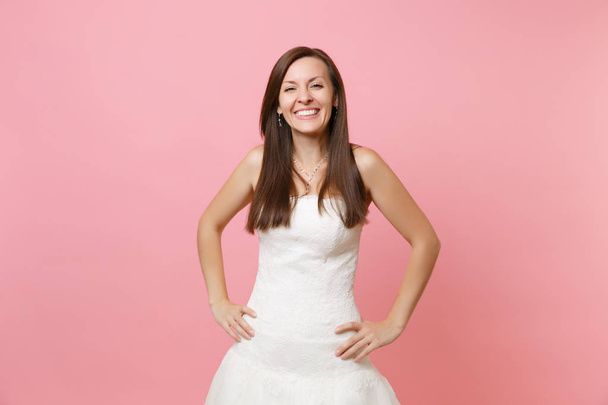Portrét radostné usměvavá nevěsta ženy v krásné bílé svatební krajkové šaty stojící s rukama v bok izolované na pastelově růžové pozadí. Svatební oslava koncept. Kopírovat prostor pro reklamu - Fotografie, Obrázek
