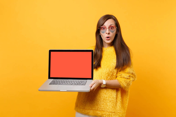 Geschokt jonge vrouw in trui, hart glazen houden pc laptop met lege zwart leeg scherm geïsoleerd op heldere gele achtergrond. Mensen oprechte emotie levensstijl concept. Gebied van de reclame - Foto, afbeelding