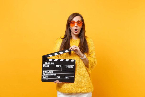 Portrait of shocked young woman in fur sweater, orange heart glasses holding classic black film making clapperboard isolated on yellow background. People sincere emotions, lifestyle. Advertising area - Photo, Image
