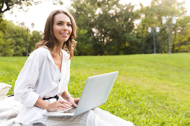 Cheerful young woman spending time at the park, sitting on a grass, using laptop computer - Фото, зображення