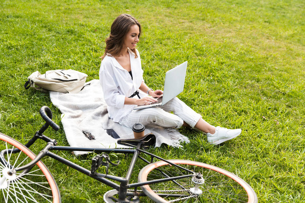 Smiling young woman bicyclist spending time at the park, sitting on a grass, using laptop computer - Foto, imagen