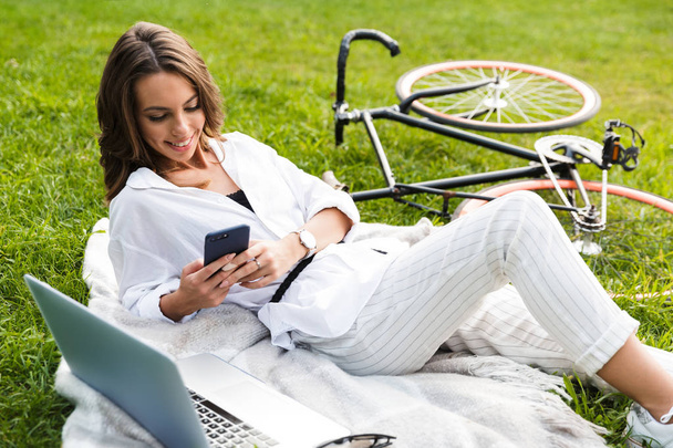 Krásná mladá žena cyklista tráví čas v parku, sedí na trávě s notebookem, držení mobilního telefonu - Fotografie, Obrázek