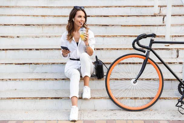 Счастливая молодая женщина проводит время в парке, сидит с велосипедом на лестнице, пьет лимонад, используя мобильный телефон
 - Фото, изображение