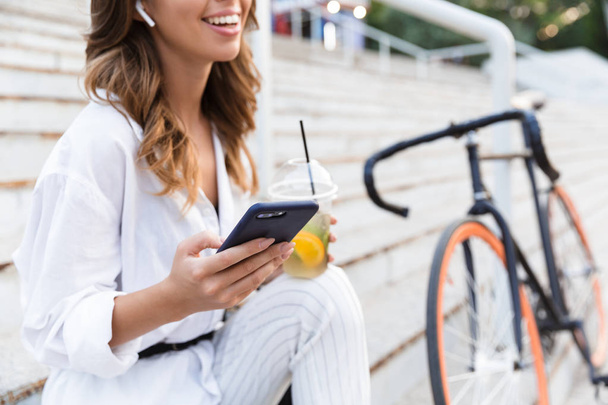 Cropped image of a laughing young woman spending time at the park, sitting with bicycle on stairs, drinking lemonade, using mobile phone - Foto, Imagen
