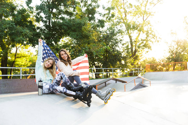 Image of happy two girls friends sisters on rollers sitting in park outdoors holding flag USA. - Φωτογραφία, εικόνα