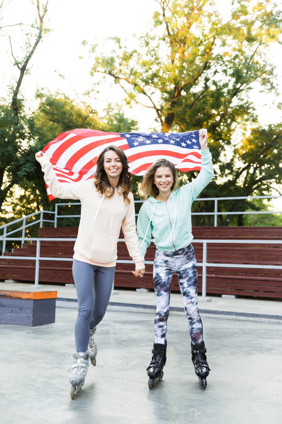 Image of happy two girls friends sisters on rollers in park outdoors holding USA flag. - Φωτογραφία, εικόνα