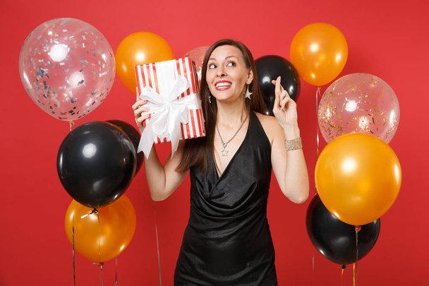 Funny young woman in black dress celebrating hold red box with gift, present keeping fingers crossed, making wish on red background air balloons. Happy New Year, birthday mockup holiday party concept - Photo, Image