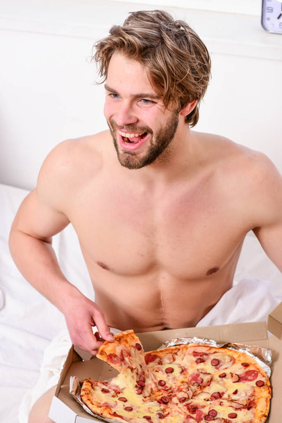 Shirtless handsome young man with pizza on bed. Guy holds pizza box sit bed in bedroom or hotel room. Portrait of lazy muscular man eating pizza while laying on a bed at home. Pizza on bed. - Photo, Image