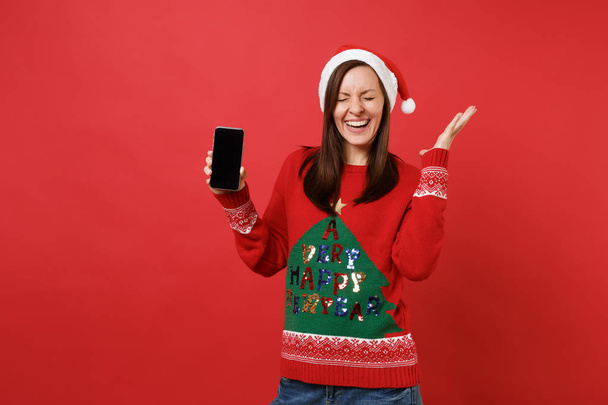 Happy Santa girl keeping eyes closed spreading hands holding mobile phone with blank empty screen isolated on red background. Happy New Year 2019 celebration holiday party concept. Mock up copy space - Фото, зображення