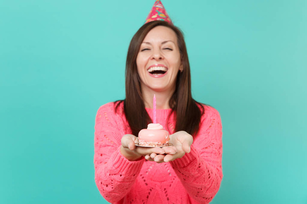Rindo jovem mulher em camisola rosa de malha e chapéu de aniversário segurando no bolo de mão com vela isolada no azul turquesa parede fundo estúdio retrato. Conceito de estilo de vida. Mock up espaço de cópia
 - Foto, Imagem