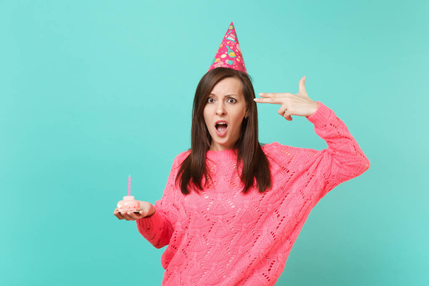 ピンクのニットのセーターの誕生日帽子の当惑の女の子は、青の背景に分離されたキャンドルとケーキを保持彼女自身撮影についてまるで頭に指をポイントします。人々 のライフ スタイルのコンセプトです。コピー スペースをモックアップします。 - 写真・画像