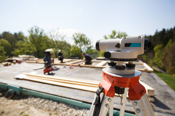 Theodolite measuring angles and positions of modules of prefabricated house. Building industry, carpentry concept with workers in the background erecting the frame. - Photo, Image