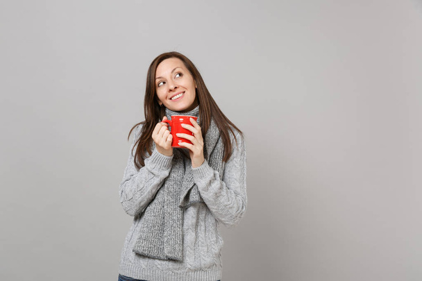 Dreamful woman in gray sweater, scarf looking up hold red cup of coffee or tea isolated on grey background. Healthy fashion lifestyle, people sincere emotions, cold season concept. Mock up copy space - Photo, Image