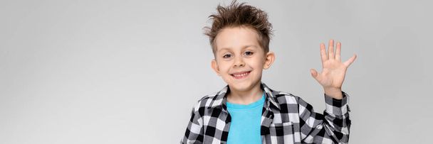 Um menino bonito em uma camisa xadrez, camisa azul e jeans fica em um fundo cinza. O menino sorri e mostra a palma da mão
 - Foto, Imagem
