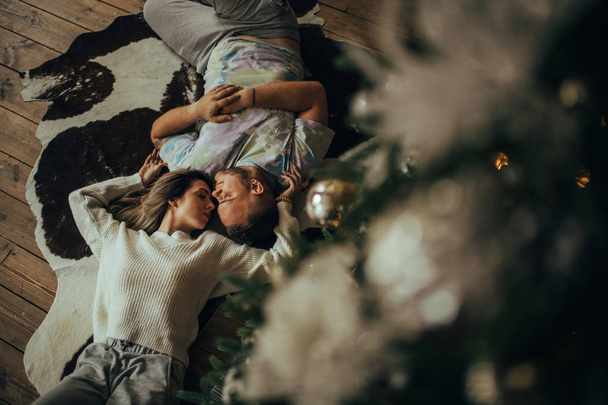 Pareja joven enamorada está acostada en la alfombra, cogida de la mano y abrazándose en el fondo del árbol de Navidad. Vista superior
. - Foto, imagen