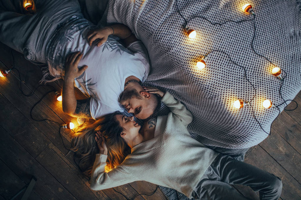 Young couple in love is lying on floor, smiling and hugging on background of glowing lightbulbs. Top view. - Fotografie, Obrázek