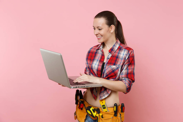 Fiatal, boldog ezermester nő felhasználó kockás ing, farmer rövidnadrág, kit eszközök öv teljes eszközök elszigetelt rózsaszín háttér laptop pc számítógépen dolgozik. Női-férfi munka. Koncepció - Fotó, kép