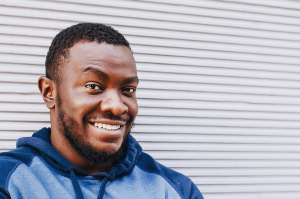 Portret van een close-up van de zwarte man in een sport trui op de achtergrond van een gestreepte lichte muur. Vrolijke positieve persoon. De foto van de stad, ruimte. Jeugdige cultuur. - Foto, afbeelding