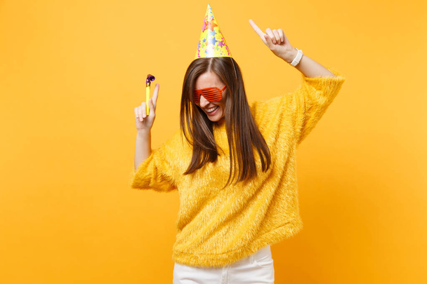 Joyful woman in orange funny glasses birthday party hat with playing pipe rising hands pointing index fingers up, dancing celebrating isolated on yellow background. People sincere emotions, lifestyle - Photo, Image