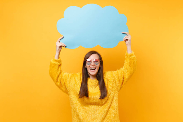 Laughing happy young woman in heart eyeglasses holding empty blank blue Say cloud, speech bubble isolated on bright yellow background. People sincere emotions, lifestyle concept. Advertising area - Foto, Bild