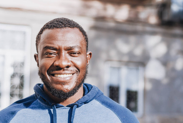 Portrait of a cheerful positive black guy close-up in a sports sweater on the background of an urban house. Race issues. City photo, space. Youthful culture. - Photo, Image
