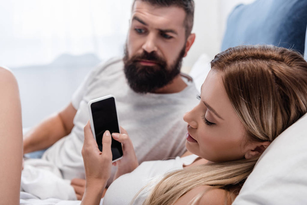 Pretty young girl lying in bed and using smartphone while bearded man looking confused - Foto, immagini
