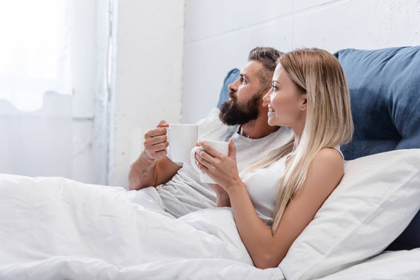 Pretty couple lying in bed and drinking coffee in bright bedroom in morning - Fotografie, Obrázek