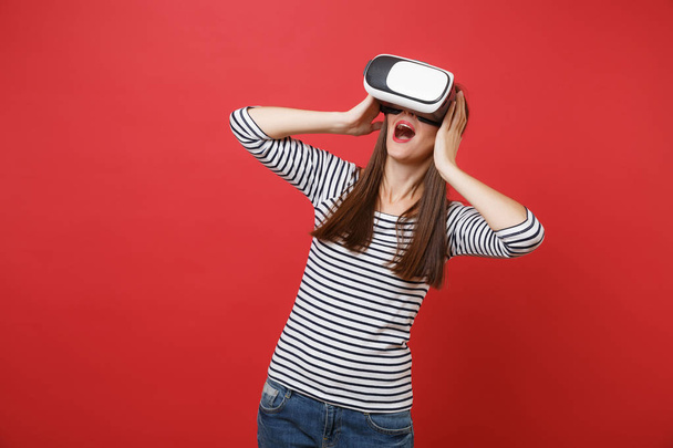 Excited young girl in striped clothes, virtual reality glasses keeping mouth wide open, putting hands on head isolated on red background. People sincere emotions lifestyle concept. Mock up copy space - Fotoğraf, Görsel