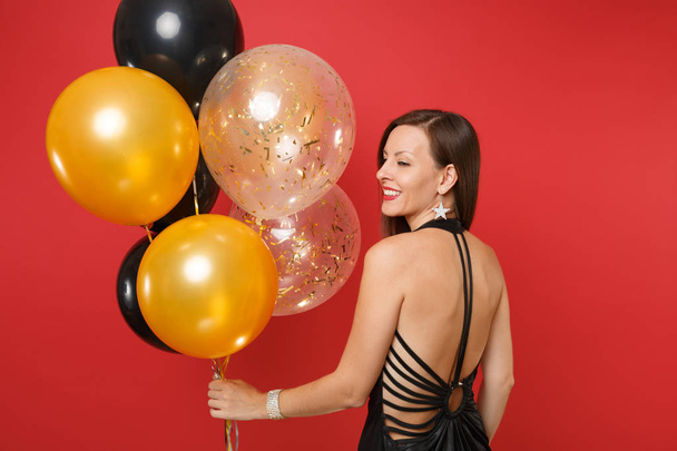Jeune femme souriante en robe noire célébrant, regardant en arrière et tenant des ballons d'air isolés sur fond rouge vif. Journée internationale de la femme Bonne année anniversaire maquette fête concept
 - Photo, image