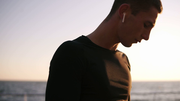 Молодой спортсмен в черной футболке надевает белые наушники и включает музыку на своем смартфоне. Слушать музыку на открытом воздухе перед морем летом
 - Кадры, видео