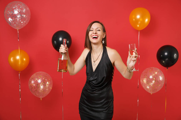 ガラス、明るい赤の背景の気球にシャンパンのボトルを保持している小さな黒いドレスを祝っての若い女性は笑ってください。女性の日、新年あけましておめでとうございます誕生日モックアップ休日党概念 - 写真・画像