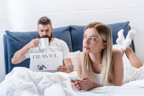jolie fille couchée et utilisant smartphone tandis que l'homme barbu boire du café et lire journal de voyage dans la chambre
 - Photo, image