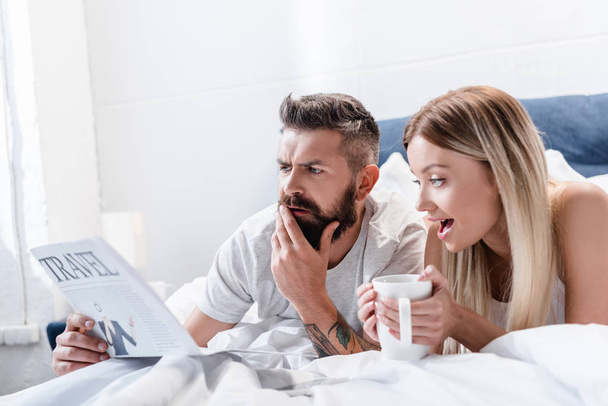 Surprised man and young woman with cup lying in bed and reading travel newspaper - Foto, afbeelding