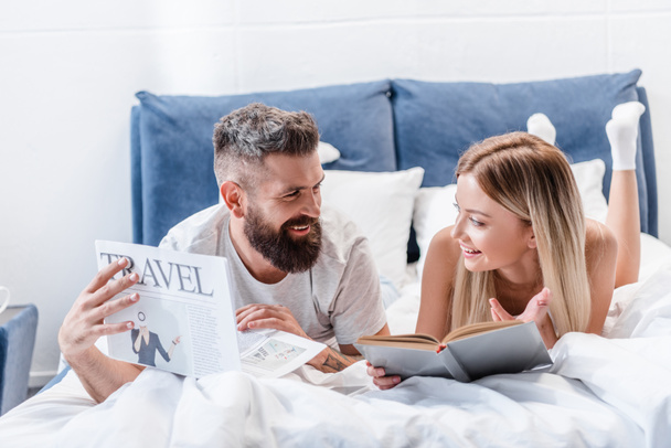 Young bearded man with travel newspaper and attractive woman with book lying in bed and looking at each other - Foto, Imagem