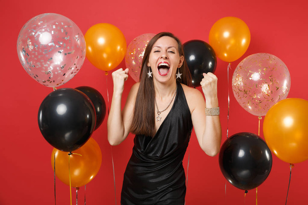 Ευτυχισμένη γυναίκα στο μαύρο φόρεμα που γιορτάζει ουρλιάζοντας, σφίγγοντας τις γροθιές σαν νικητής στο λαμπερό κόκκινο φόντο και αερόστατα. Διεθνές Γυναικείο ημέρα, ευτυχισμένο το νέο έτος, γενέθλια mockup διακοπών κόμμα έννοια - Φωτογραφία, εικόνα