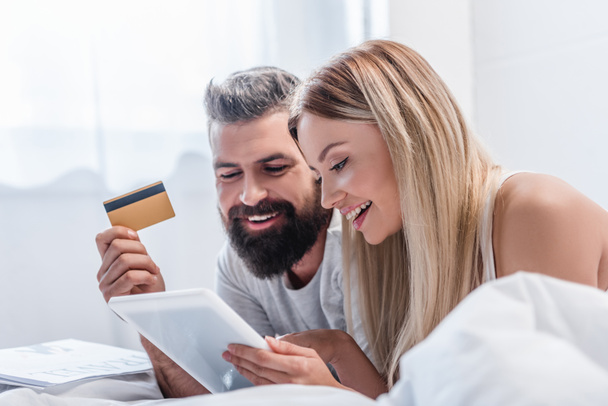 Atractiva pareja con tableta digital y tarjeta de crédito en el dormitorio luminoso
 - Foto, imagen