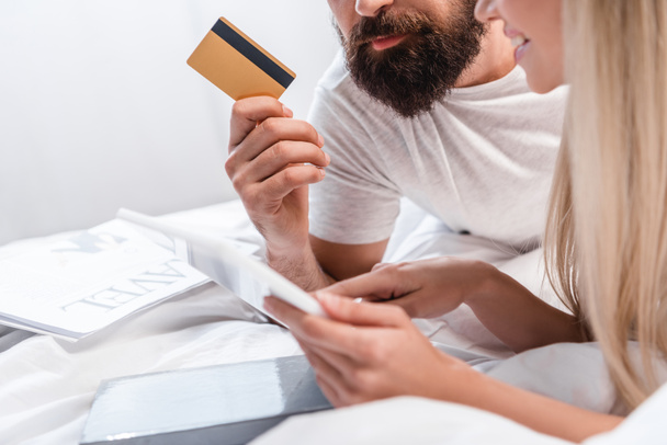 Young woman pointing with finger at digital tablet while bearded man holding credit card in bedroom - Fotó, kép