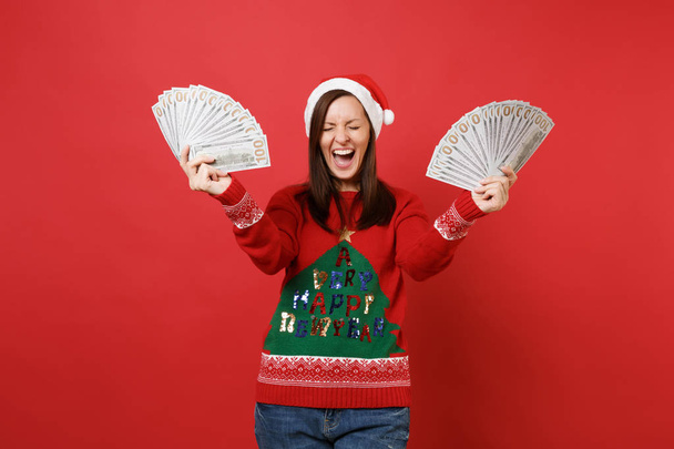 Счастливая Санта девушка с закрытыми глазами кричит, держа много банкнот долларов, наличные деньги изолированы на красном фоне. Концепция праздничной вечеринки в честь Нового 2019 года. Пространство для копирования
 - Фото, изображение