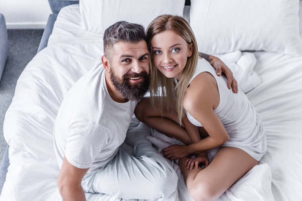 overhead view of happy young couple sitting in white bed and looking at camera - Valokuva, kuva