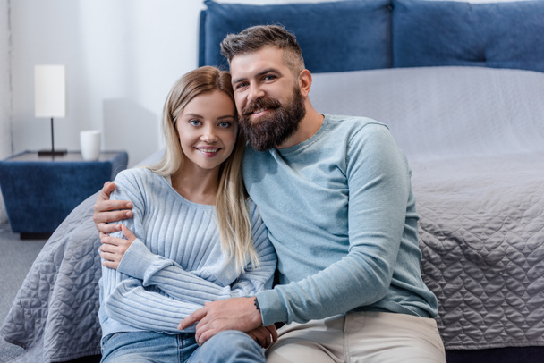 happy young couple sitting on floor, smiling and looking at camera in blue bedroom - Foto, Imagen
