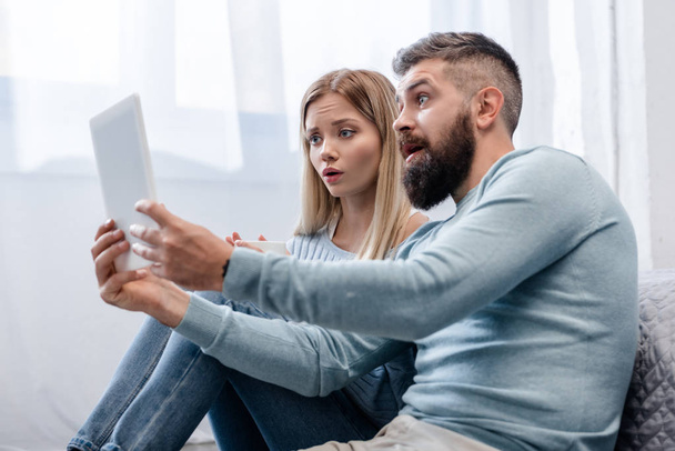Couple sitting on floor with digital tablet and looking surprised - Foto, afbeelding