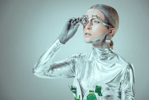 futuristic silver robot adjusting eye prosthesis and looking away isolated on grey, future technology concept   - 写真・画像