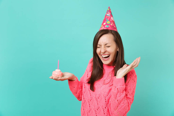 Jonge vrouw lachen in gebreide roze trui, verjaardag hoed hold girosaldi taart met kaars, verspreiden handen, ogen gesloten houden geïsoleerd op blauwe achtergrond. Mensen levensstijl concept. Bespotten kopie databaseruimte - Foto, afbeelding