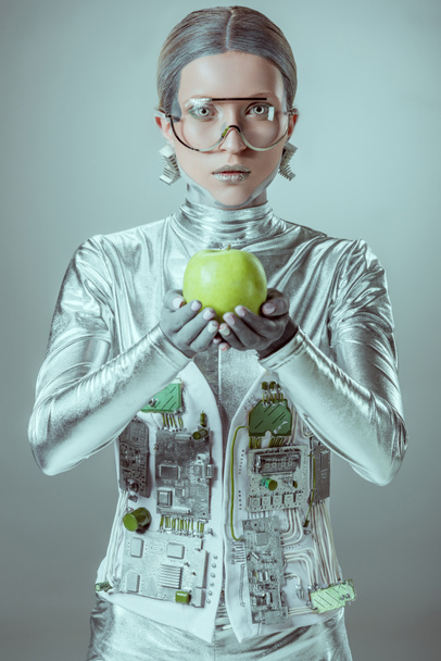 cyborg in futuristic eyeglasses holding apple and looking at camera isolated on grey, future technology concept  - Valokuva, kuva