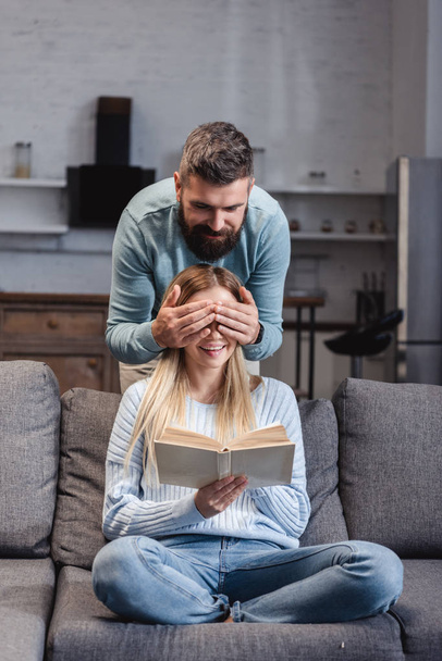 Cheerful husband closing eyes of happy wife with book  - Photo, image