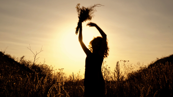 Czarne sylwetki dziewczyny na tle zachodu słońca jasny. Ona skacze i fale jej ręce wśród kwiatów w polu między wzgórzami. Widok z dołu. Z bliska. 4 k. 25 fps. - Materiał filmowy, wideo